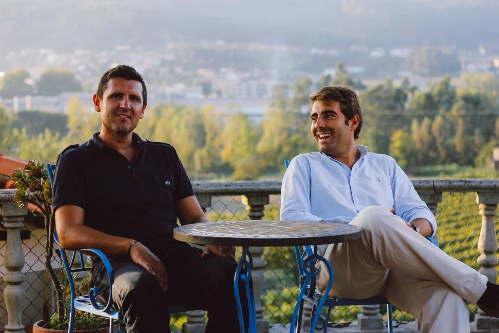 Dois homens sentados em uma varanda com vista para uma cidade e árvores, sorrindo e conversando.