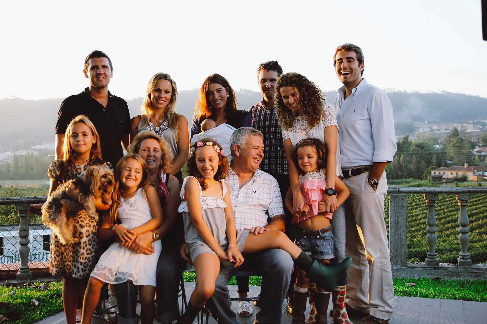 Um grupo de pessoas sorrindo em um encontro ao ar livre durante o pôr do sol, incluindo adultos, crianças e um cachorro, com fundo de montanhas e céu claro.