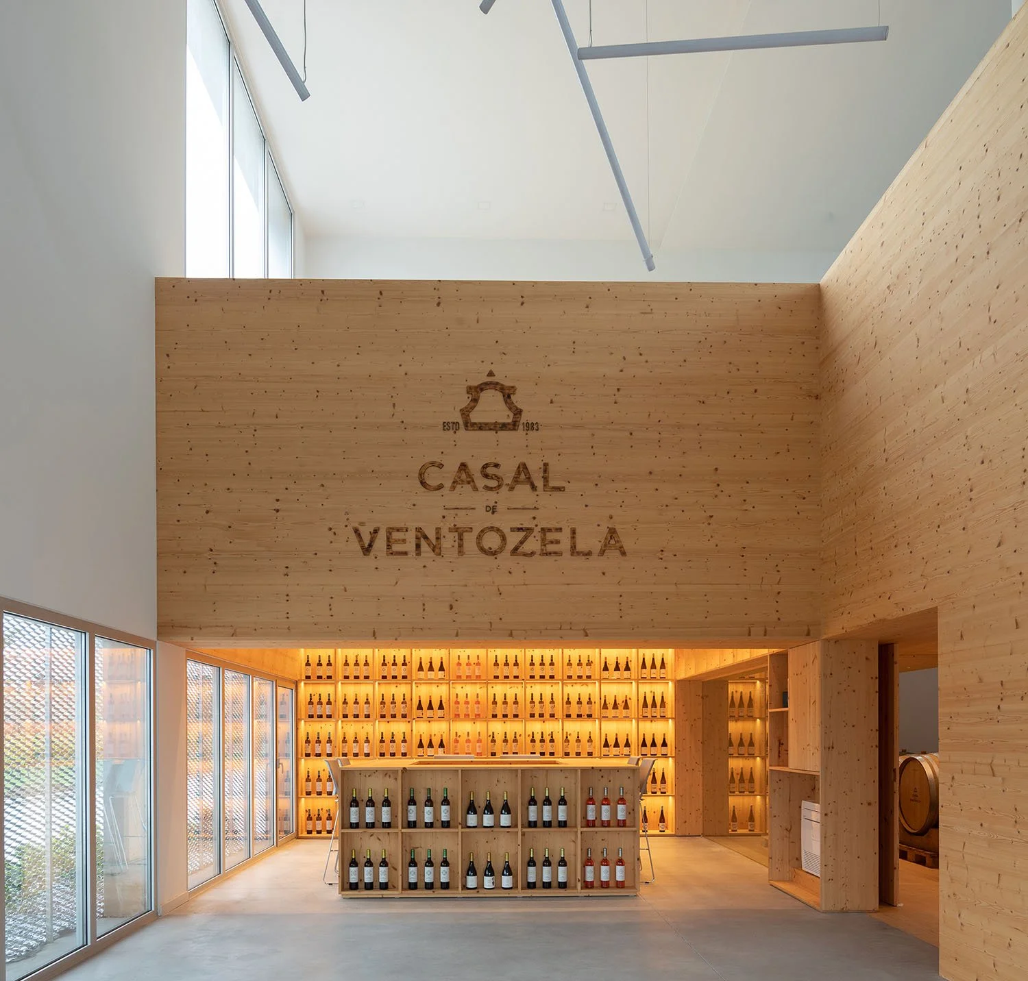 Interior de uma loja de vinhos com paredes de madeira, prateleiras cheias de garrafas de vinho, iluminação quente e janelas de vidro.