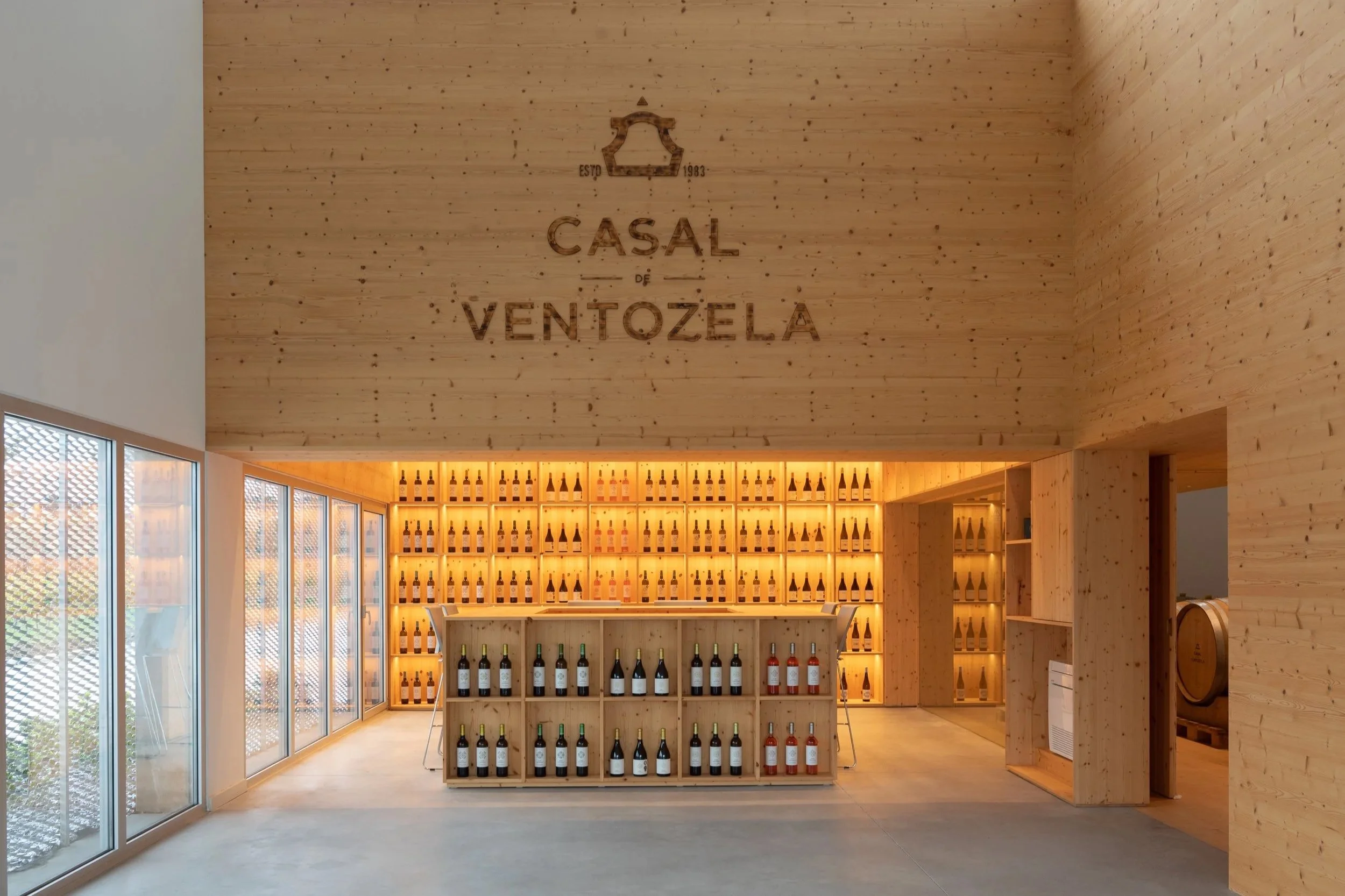 Interior de uma loja de vinhos com paredes de madeira, prateleiras cheias de garrafas de vinho, iluminação quente e janelas de vidro.