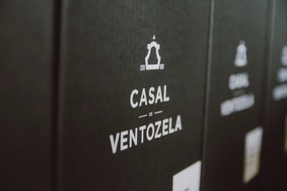 Caixas pretas com etiquetas brancas, uma delas com o texto 'Casal de Ventozela' e símbolo de uma torre no topo, indicando uma embalagem de vinho.