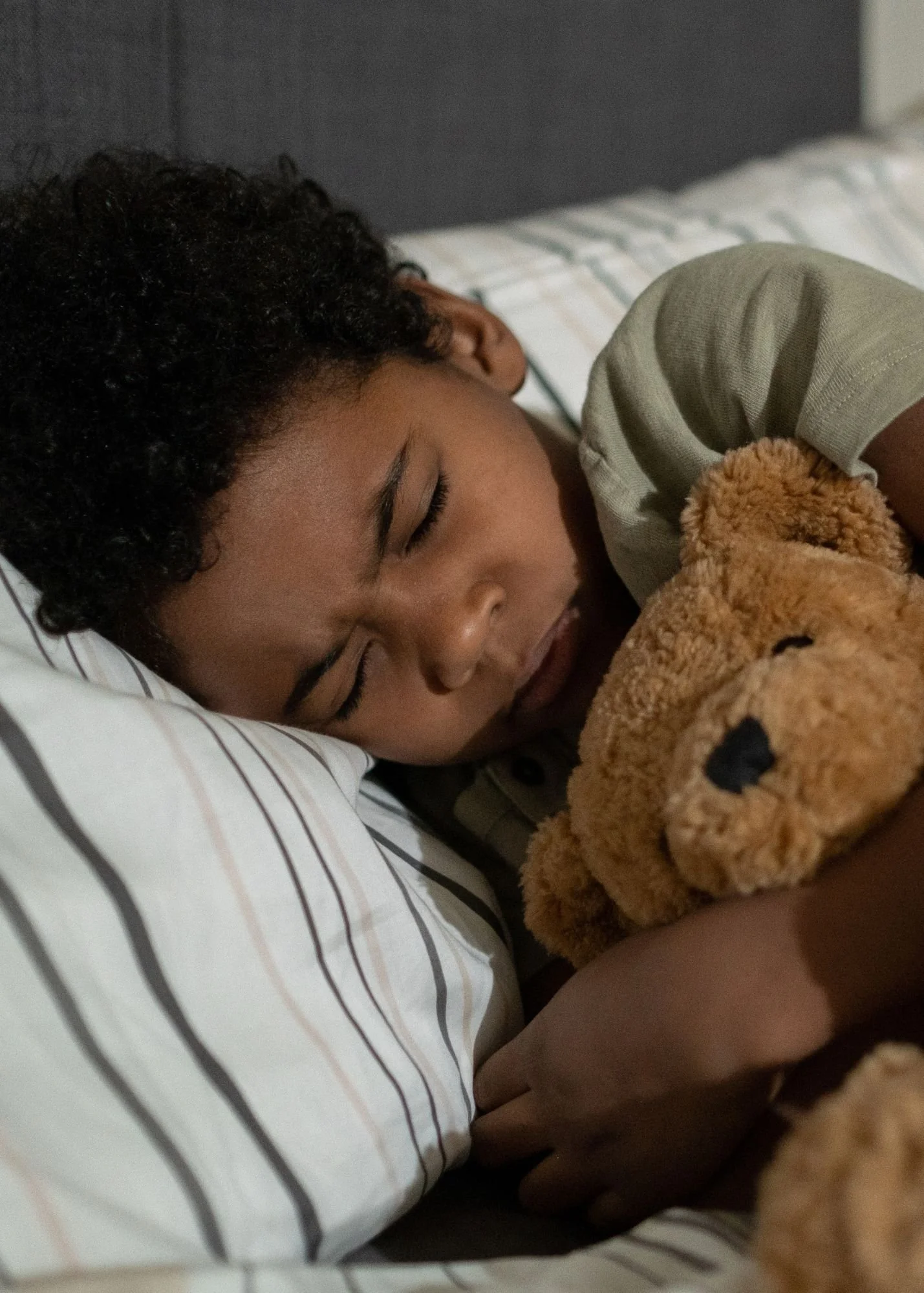 A young boy with curly hair sleeping peacefully on a pillow, hugging a brown teddy bear, in a bed with white striped sheets.
