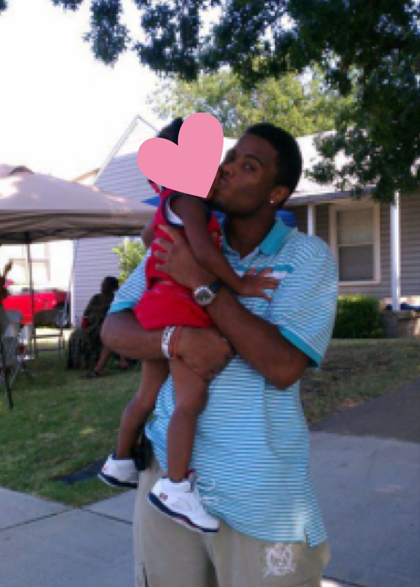 A man is holding a child in his arms outdoors in a neighborhood. The man is wearing a light blue and white striped shirt, tan shorts, and a watch. The child is wearing a red and white striped shirt and red shorts, with a pink heart covering the child's face. The background shows houses, green trees, and a fenced yard.