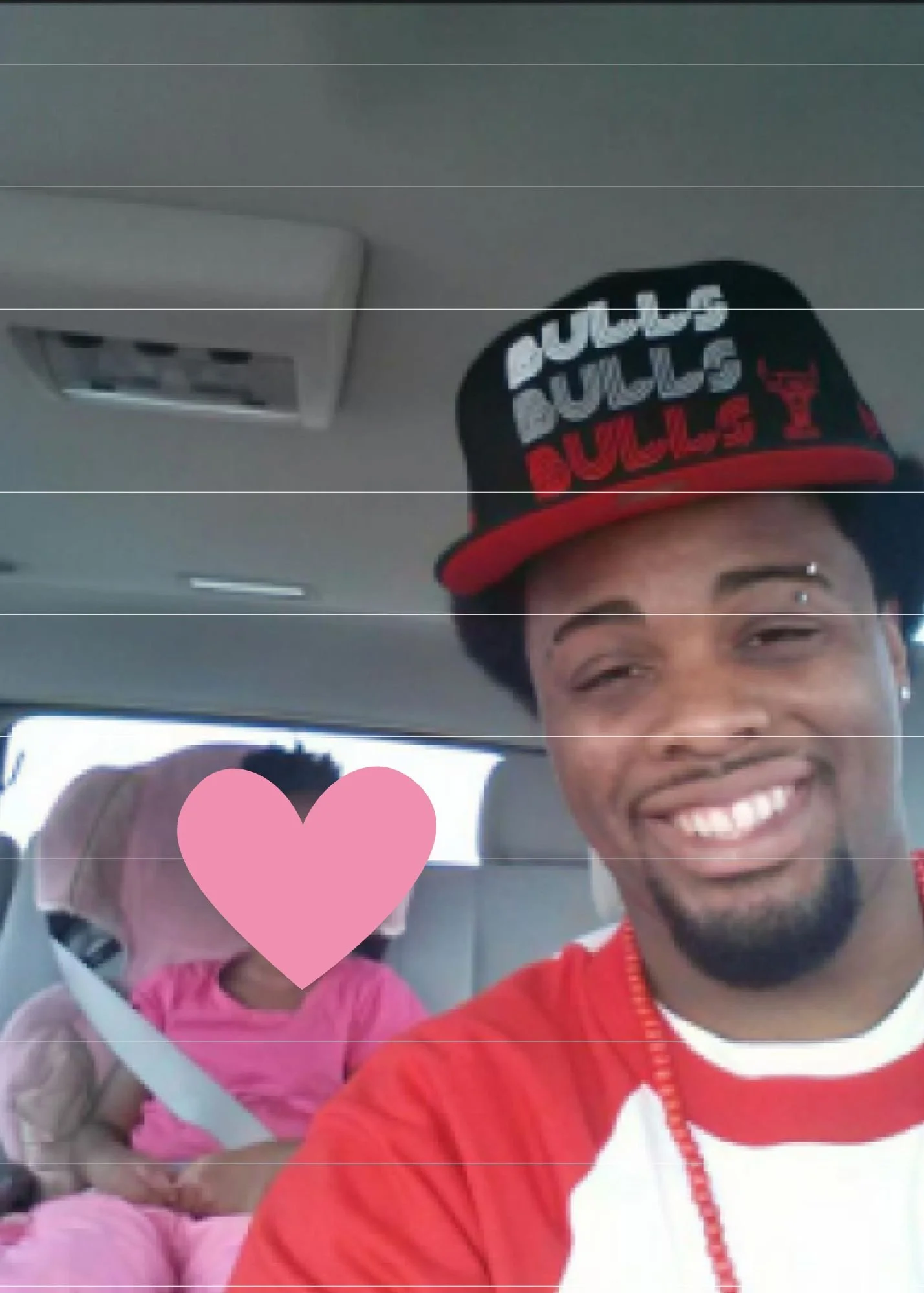 Amos Wells, a man smiling in a red and white jacket taking a selfie with a woman wearing a pink hoodie and a pink heart over her face, sitting in a vehicle.