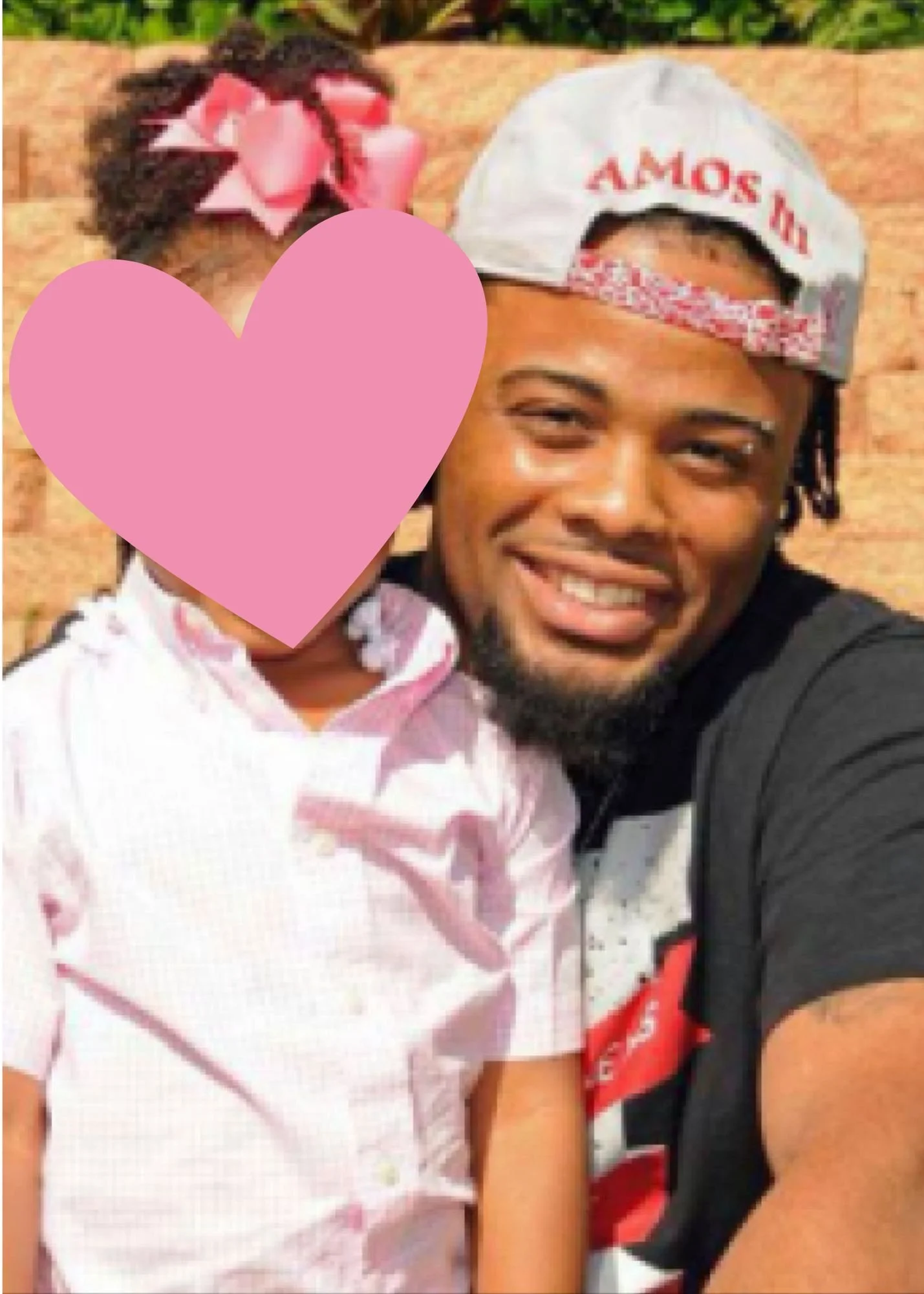 Amos Wells smiling with a beard wearing a gray cap and black T-shirt taking a selfie with his daughter as a girl in a white and pink uniform. The girl's face is covered with a pink heart shape. The background shows a brick wall and some greenery.