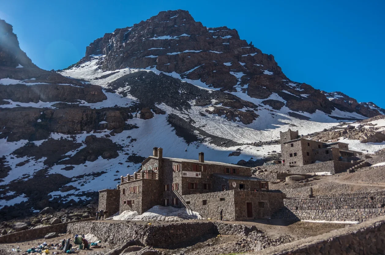 A mountain landscape with snow patches, featuring stone buildings at the base, some with chimneys, under a clear blue sky.