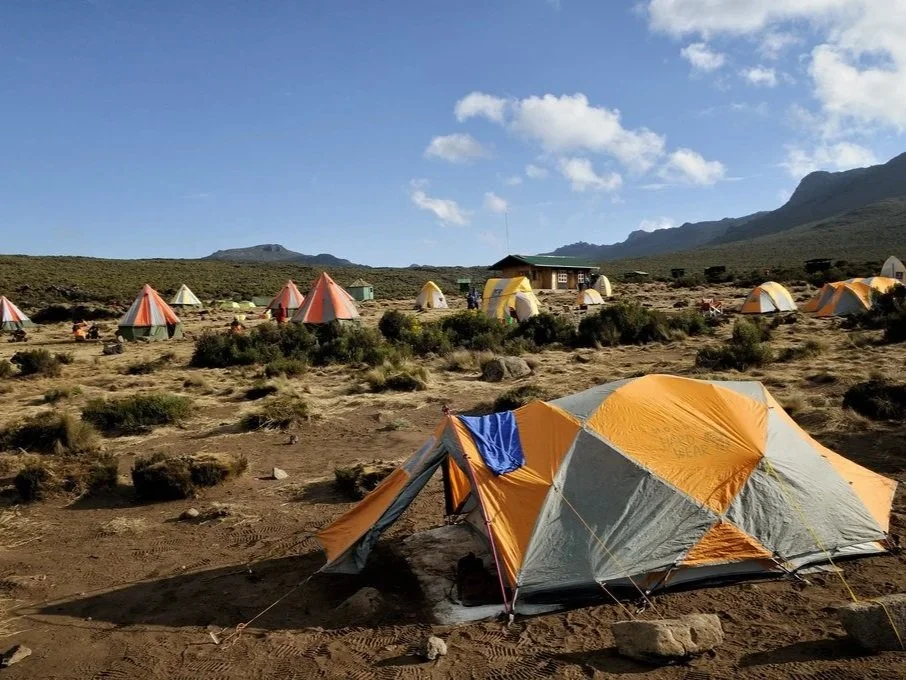 Multiple tents set up in an open, rocky landscape with sparse vegetation, mountains in the background, and a blue sky with some clouds.