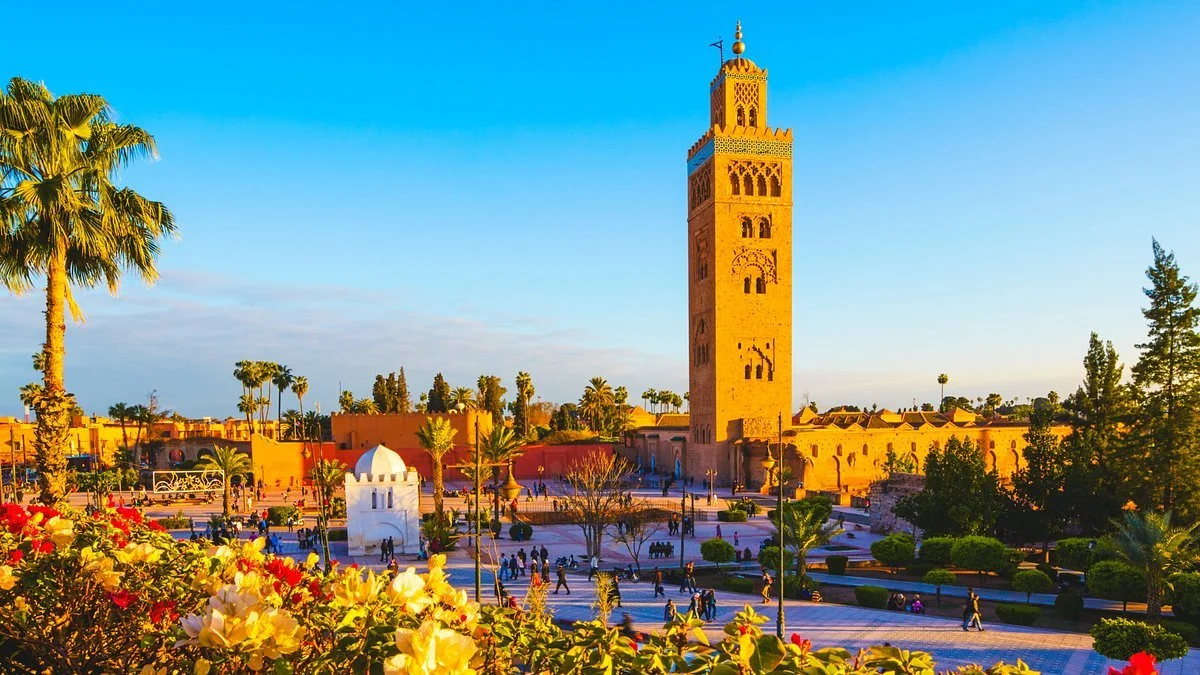 A scenic view of the Moroccan Koutoubia Mosque and its tall minaret, surrounded by palm trees, flowering bushes, and people walking in a plaza during daytime with a clear blue sky.