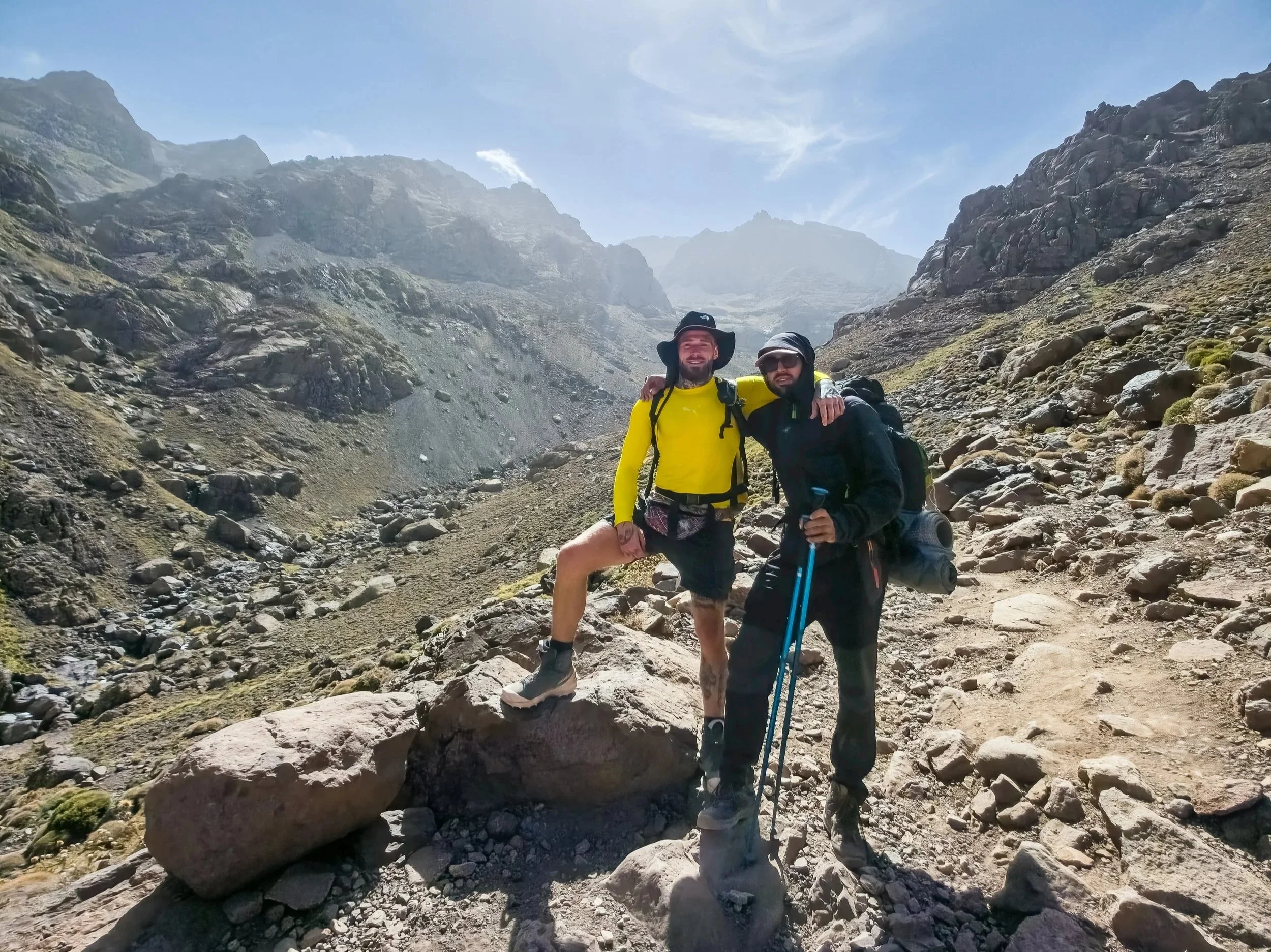 Two men standing together outdoors in a rocky mountain landscape, embracing and smiling. One is wearing a yellow long sleeve shirt and a black bucket hat, the other is wearing black clothing, a black hat, sunglasses, and holding a trekking pole.