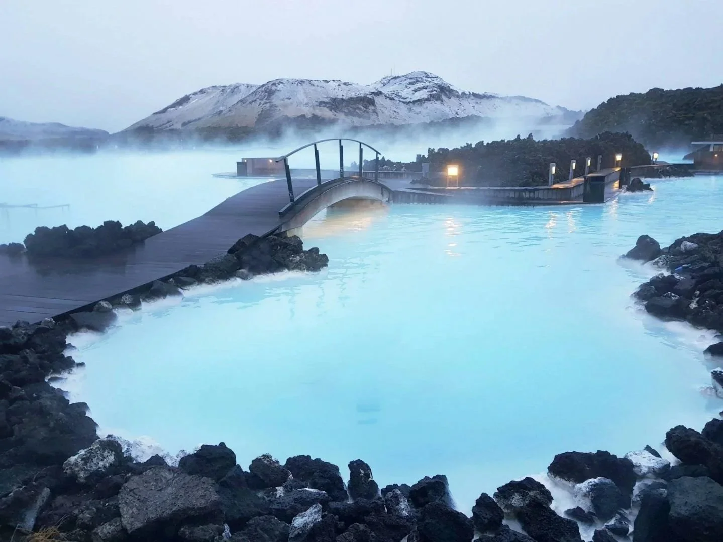 A hot spring pool with rocky edges, a wooden bridge, and a mountain in the background, surrounded by steam and mist.
