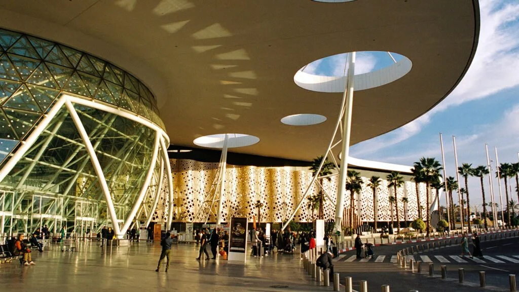 Modern building with glass and steel architecture, palm trees, and people walking outside on a sunny day.