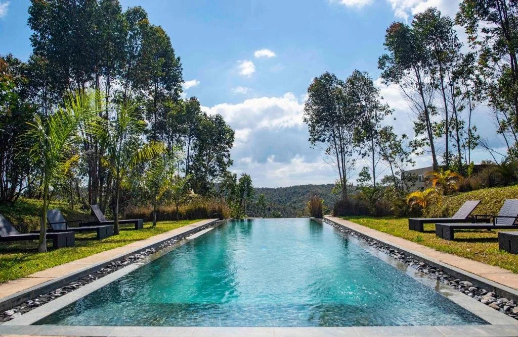 A rectangular swimming pool with lounge chairs on both sides, surrounded by trees and greenery, under a partly cloudy sky.