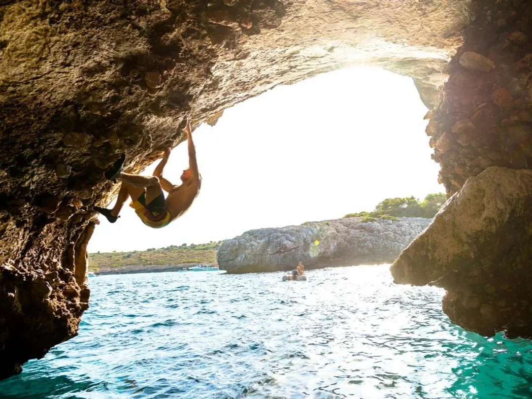 A person rock climbing on a cliff near water, with another person swimming in the water below, and rocky formations in the background.