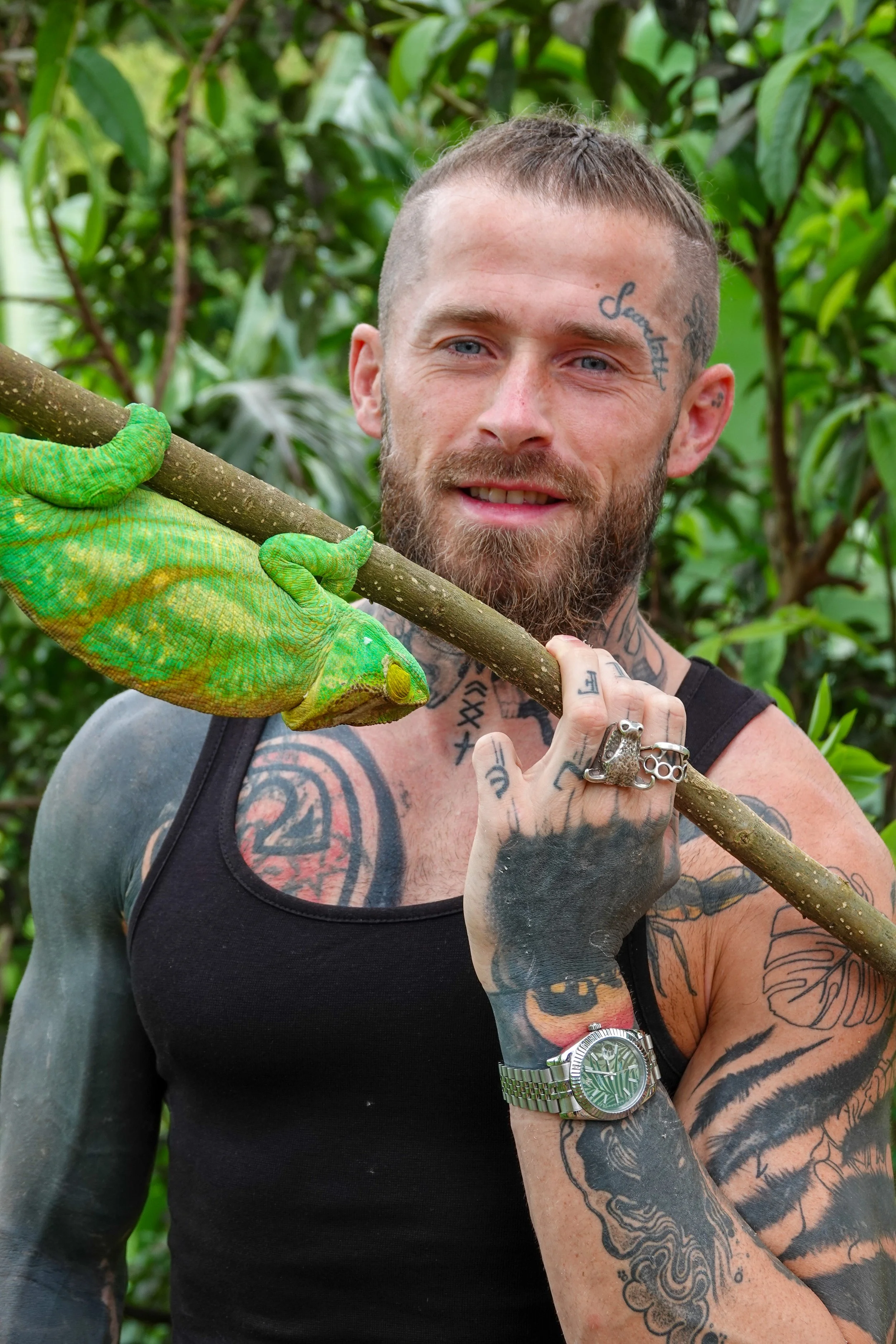 A man with tattoos and a beard holding a large branch with green gloves in a lush, green environment.