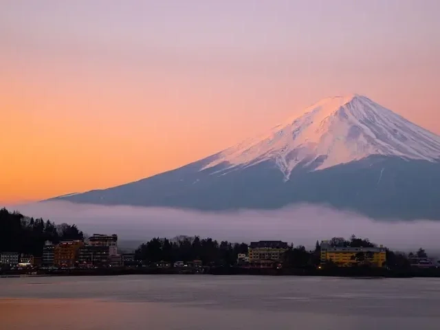 Scenic view of Mount Fuji at sunrise, with city buildings and a body of water in the foreground.
