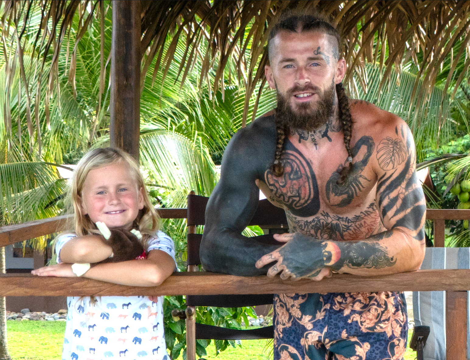 A young girl with blonde hair and a man with tattoos, long hair in braids, and a beard, both leaning on a wooden railing outdoors with green tropical plants in the background.