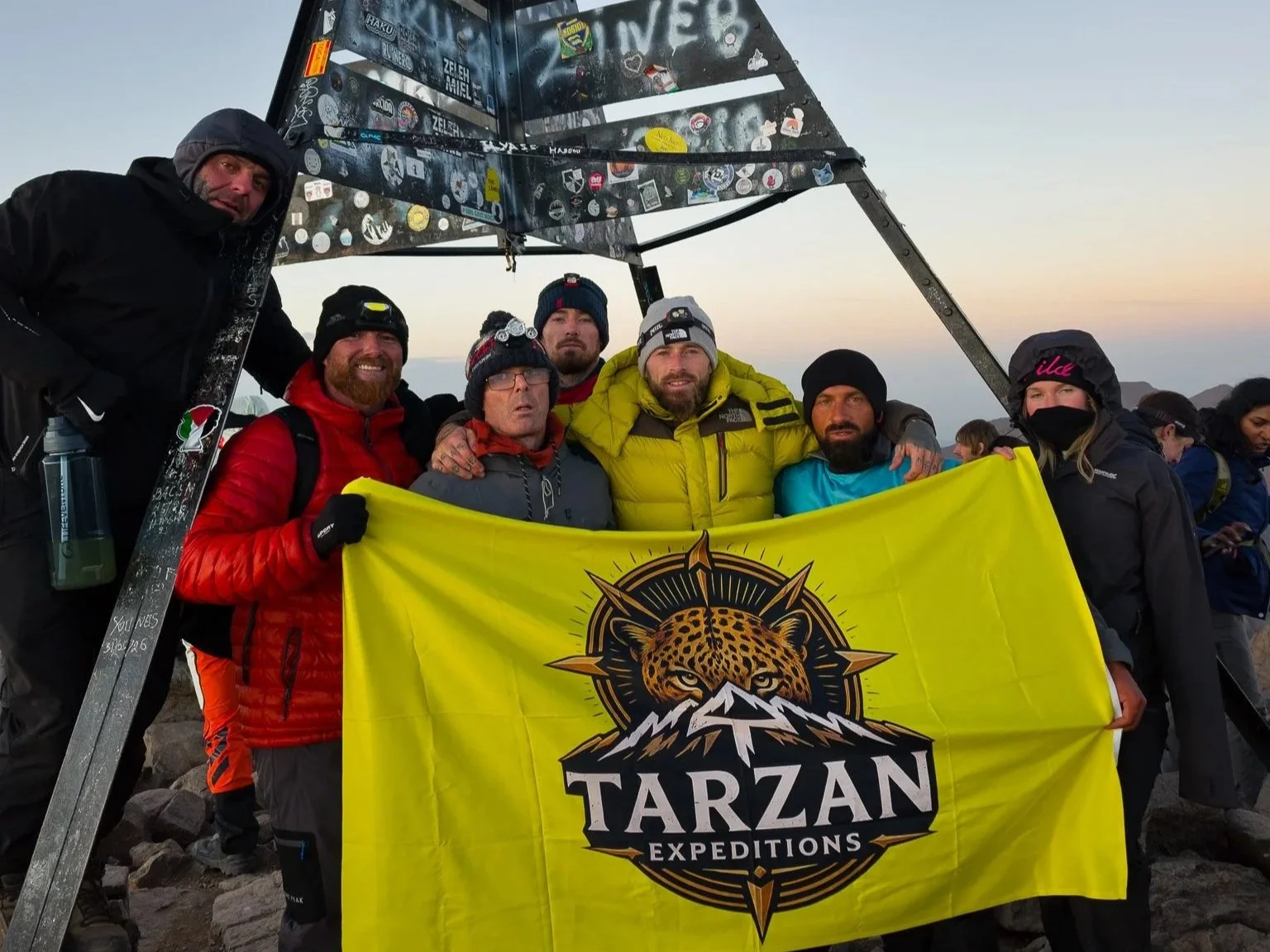 Group of seven climbers on the summit of Mount Toubkal holding a yellow 'Tarzan Expeditions' flag, with a pyramid-shaped structure covered in stickers behind them at sunrise.