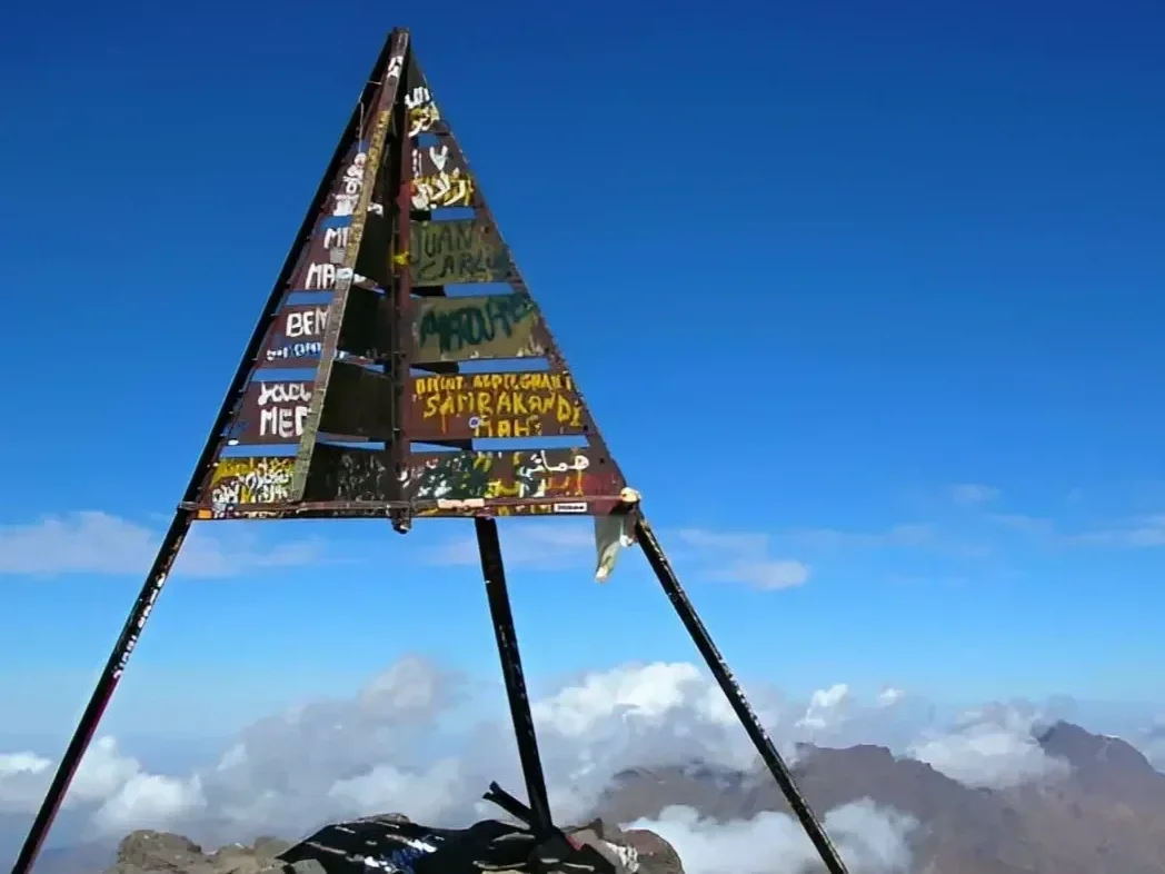 A triangular metal structure with painted words and symbols on a mountain summit, with a clear blue sky and clouds below.