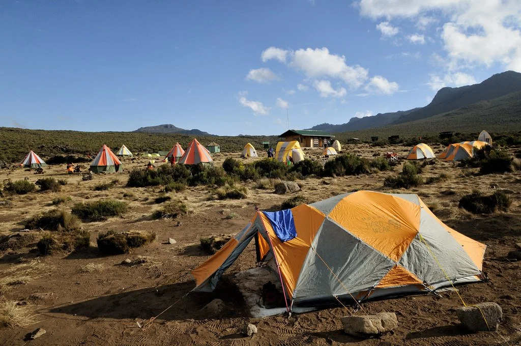 A desert campsite with multiple orange and yellow tents, some with striped patterns, set up on sandy ground with sparse vegetation. There is a small building and mountains in the background under a partly cloudy sky.