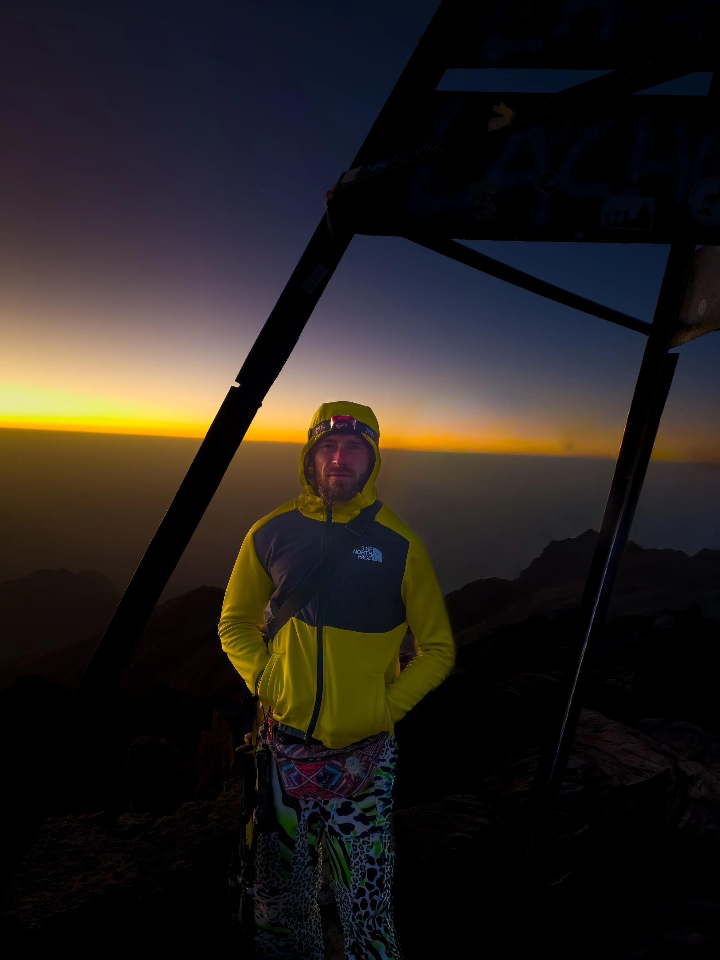 A man wearing a yellow jacket and patterned pants stands on a mountain at sunset with a paraglider canopy above him.