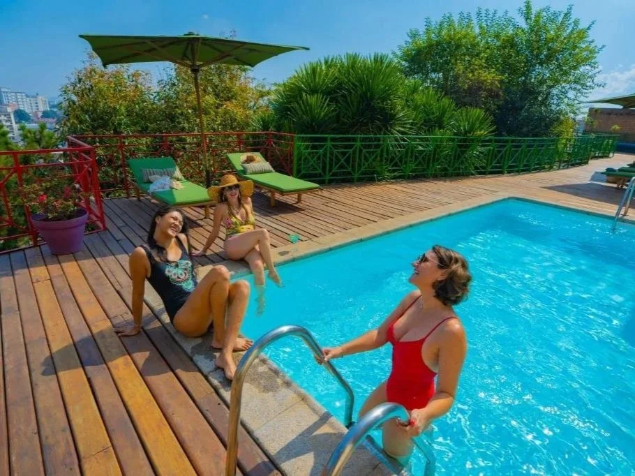 Three women enjoying a sunny day by a swimming pool on a wooden deck, with two lounging chairs, an umbrella, and lush greenery in the background.