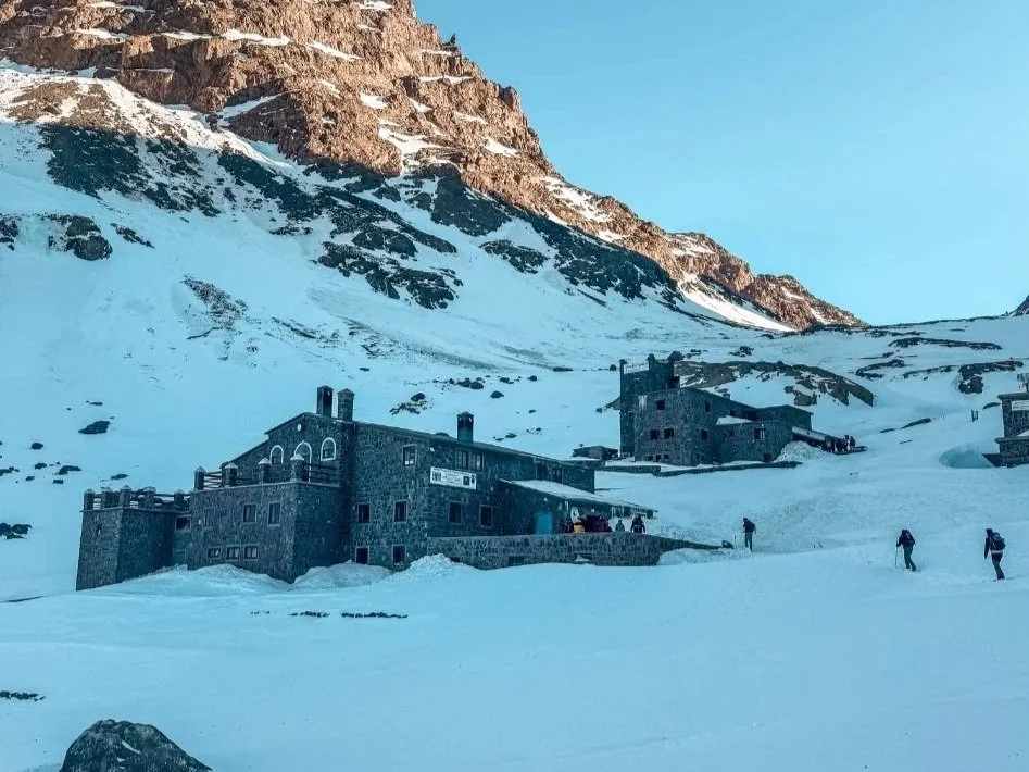 Snow-covered landscape with dark stone buildings at the base of mountain slopes, some people walking in the snow.