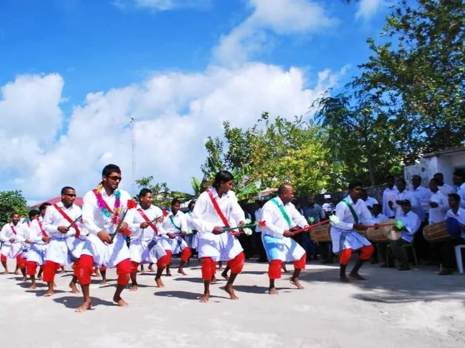A group of men dancing outdoors, wearing white traditional attire with red sashes and pants, holding sticks, with a crowd of onlookers and trees under a partly cloudy sky.