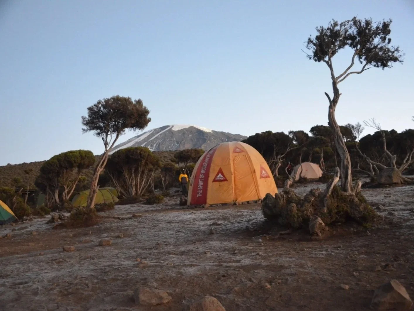 Camping tents set up in a rocky, arid landscape with sparse trees, mountains in the background, and snow on a distant mountain peak.