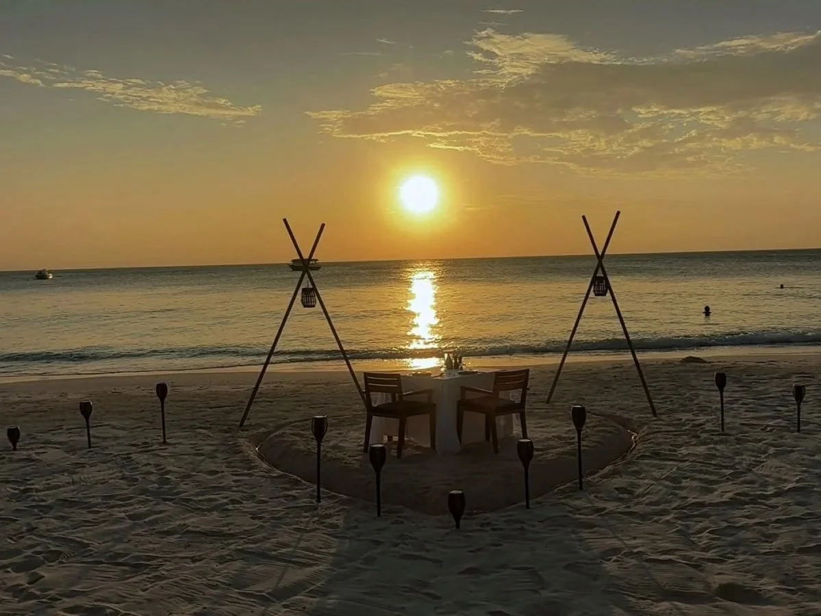 Set up for a beach wedding with a table and chairs facing the sunset, two teepees, and tiki torches on the sand shoreline during sunset.