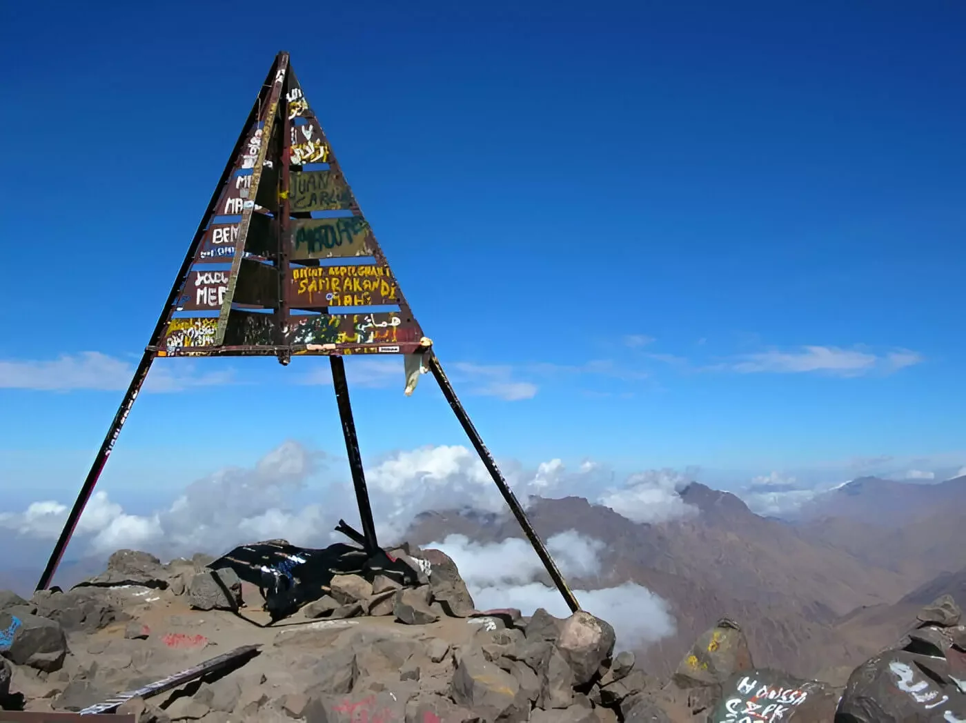 A triangular metal sign on a rocky mountain summit with graffiti, surrounded by clouds and distant mountain peaks under a clear blue sky.