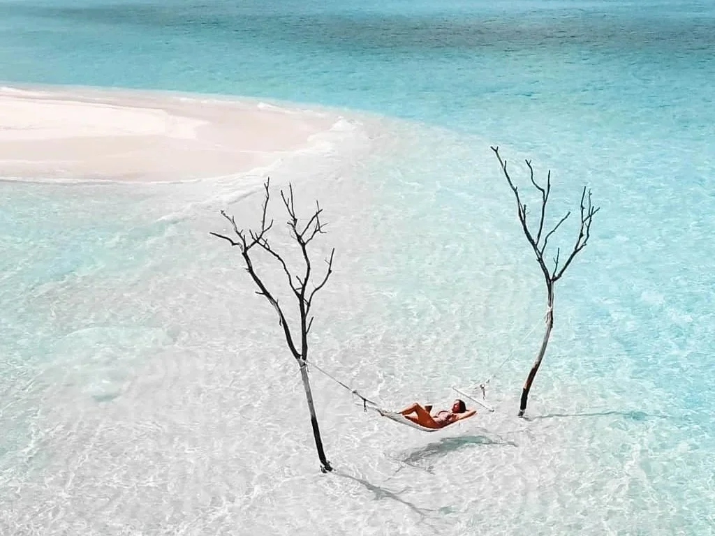 A person lying in a hammock tied between two leafless trees on a white sandy beach with turquoise water in the background.