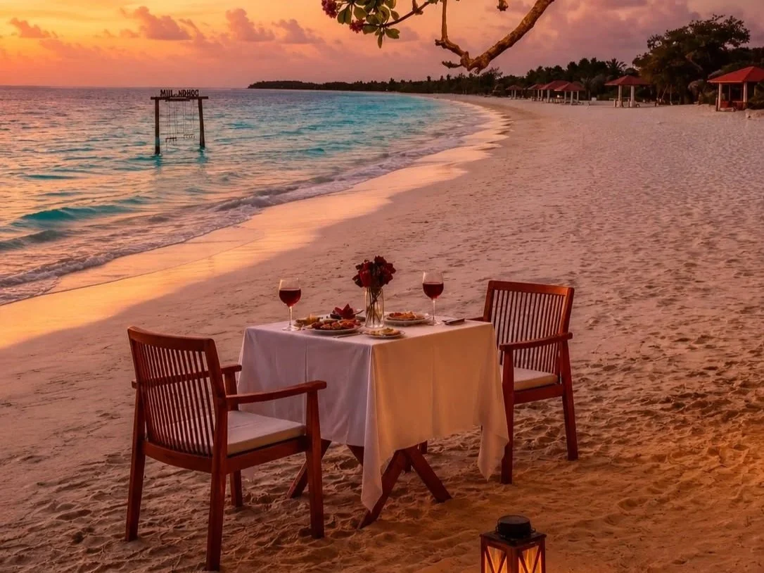 A romantic beach dinner setup on the sand with a table, two chairs, a vase of flowers, and two glasses of red wine, overlooking an ocean at sunset with colorful sky and distant huts.