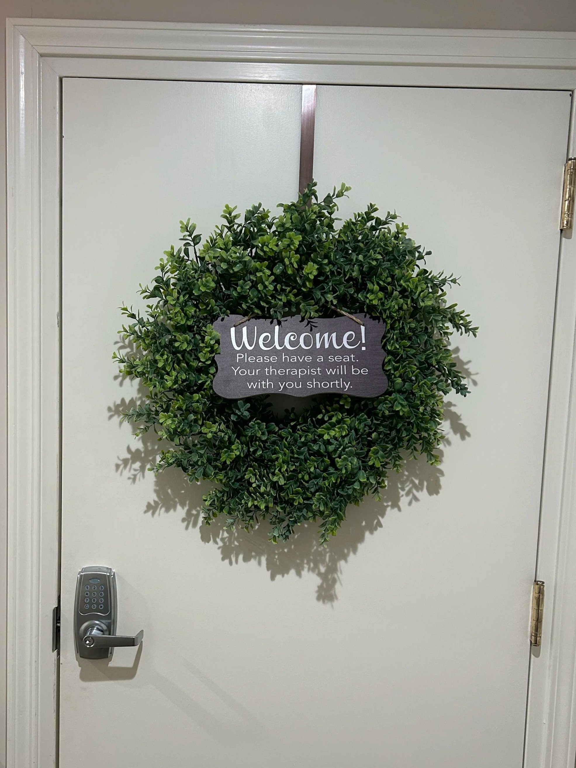 Front door decorated with a green wreath and a black sign that says 'Welcome! Please have a seat. Your therapist will be with you shortly.'