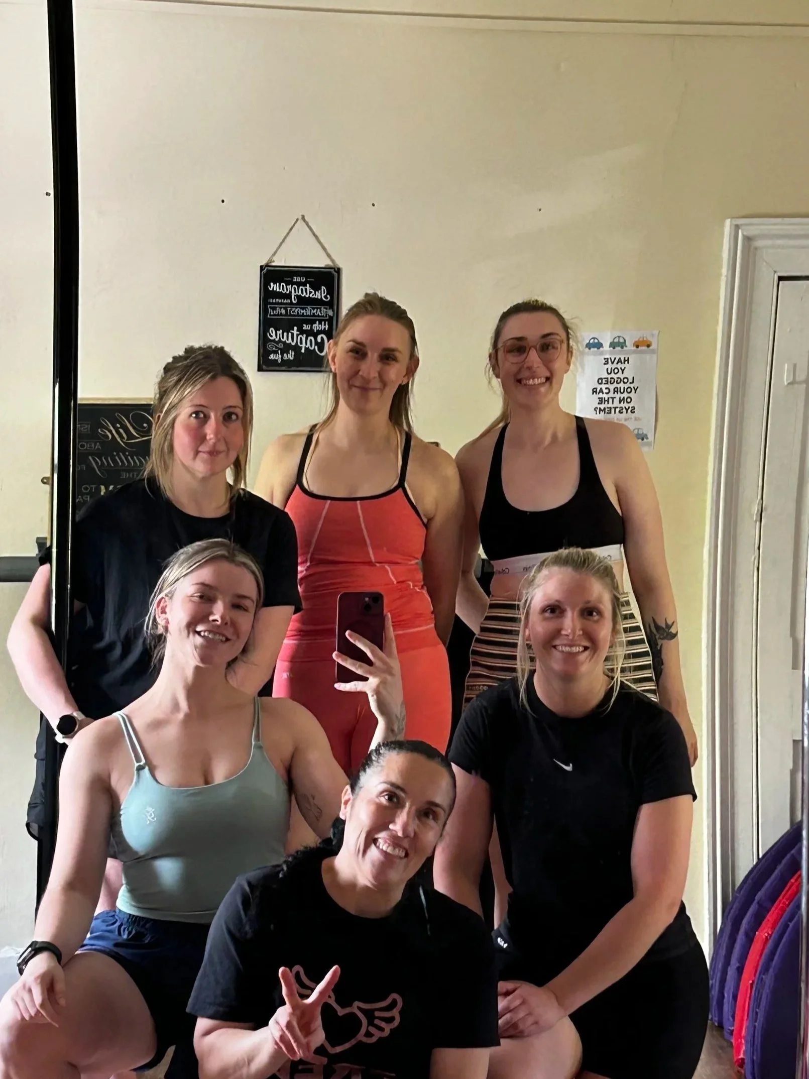 Group of people taking a mirror selfie in workout attire, standing in a gym or fitness studio, with colorful equipment hanging on the side and motivational signs on the wall.