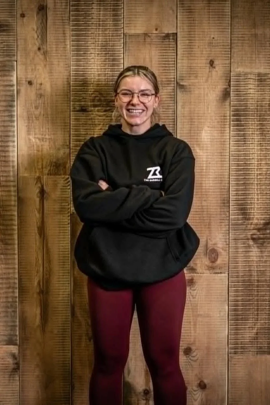 A woman with glasses, smiling, wearing a black hoodie with logo, and maroon leggings, standing in front of a wooden wall.