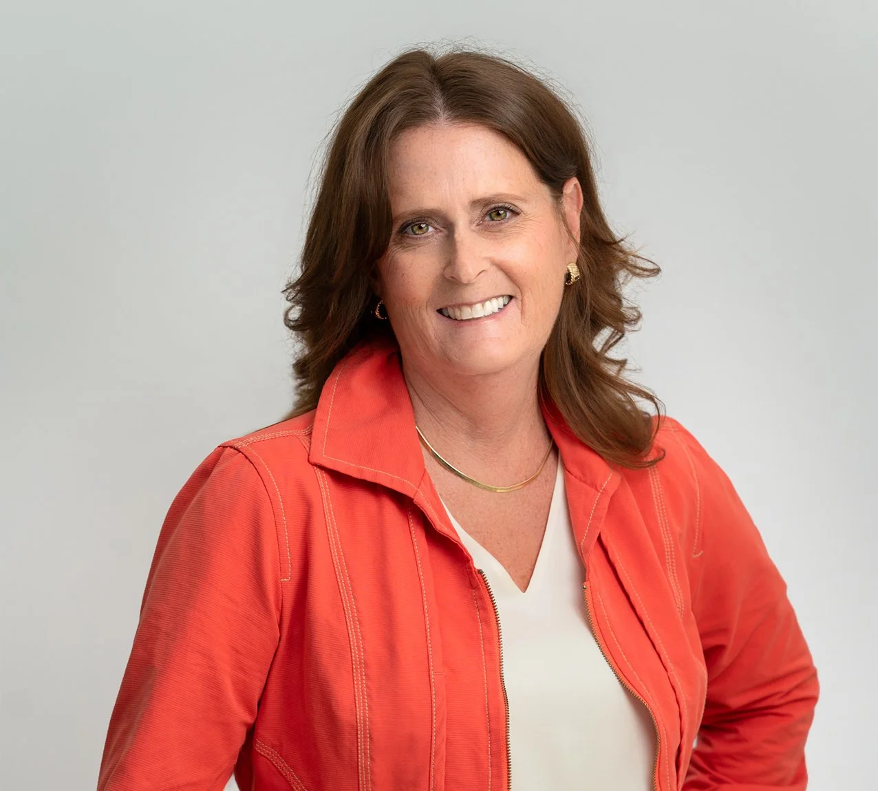 Angie Clark Consulting | Human + Resources. Smiling, wearing a coral jacket over a white top, gold necklace, and earrings, standing against a light grey background.