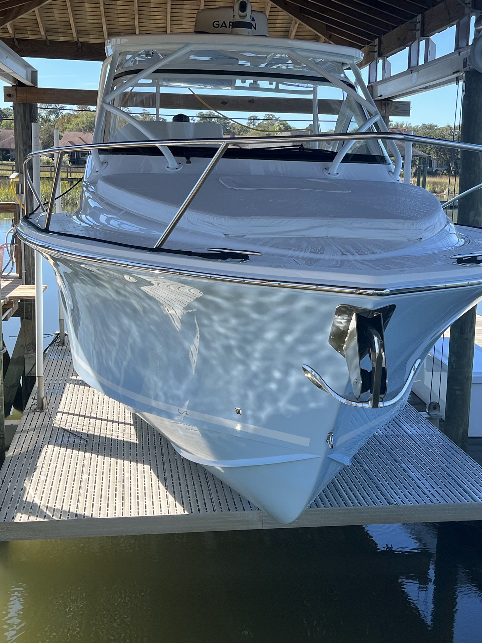 White motorboat docked at a covered marina with a view of houses and trees in the background under a clear sky.