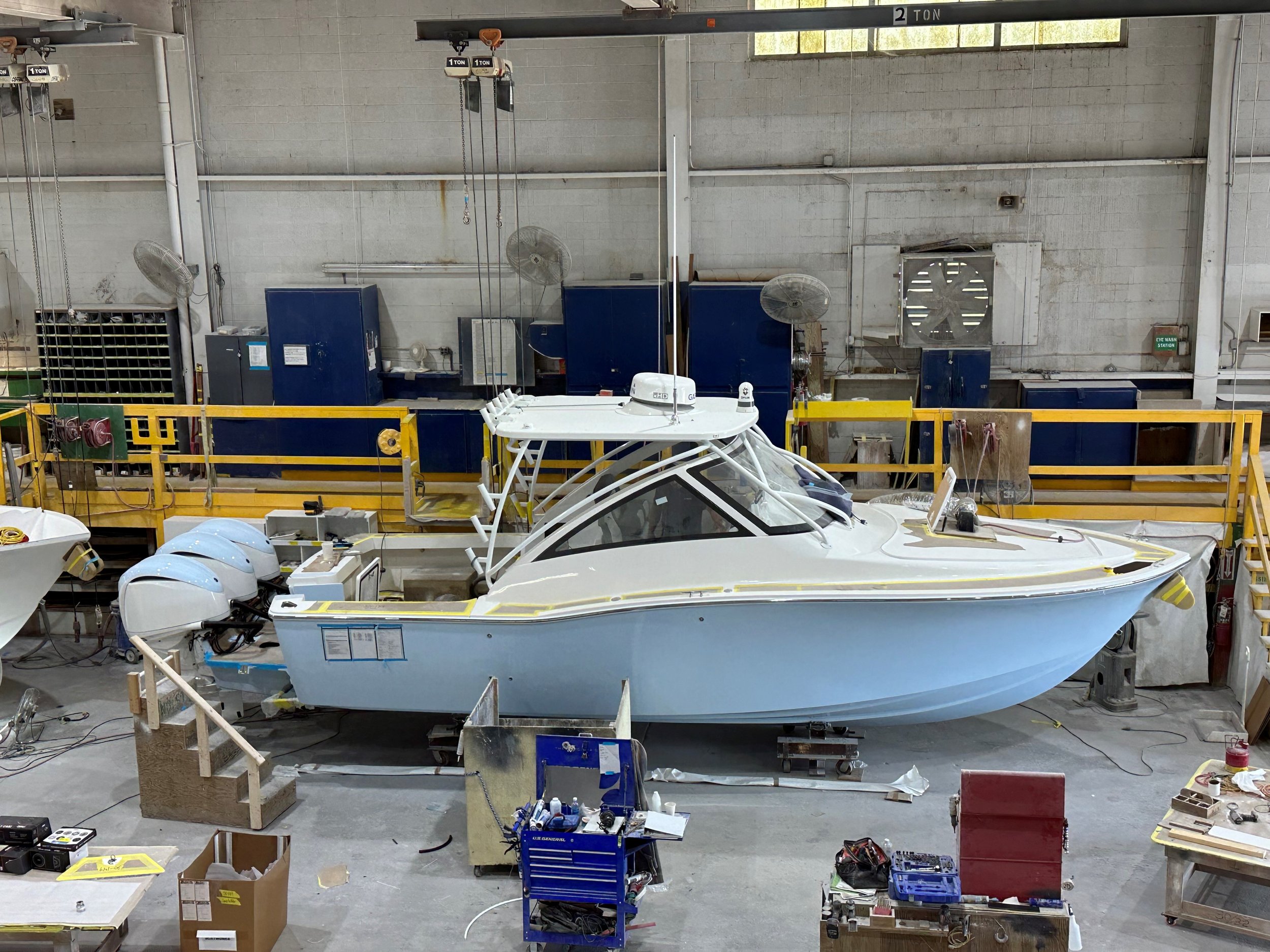 A white yacht in a boat manufacturing or repair facility, surrounded by tools, equipment, and workbenches with shelves. The background features a gray cinder block wall with industrial fans and electrical components.