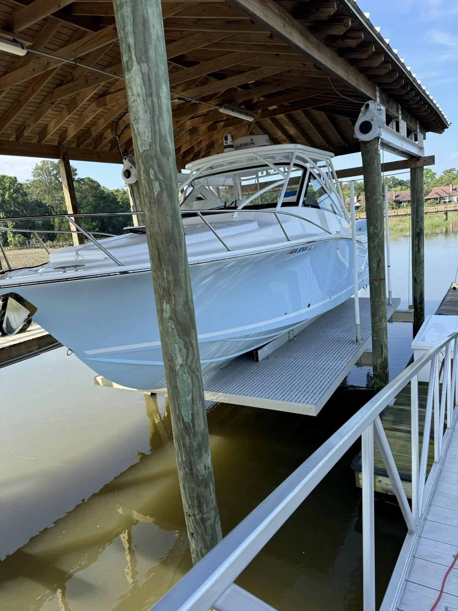 A white motorboat docked in a boat lift at a marina, with a wooden roof structure overhead and water underneath. The boat has a Garmin radar on top and is secured on a metal platform with surrounding water and a partly cloudy sky.
