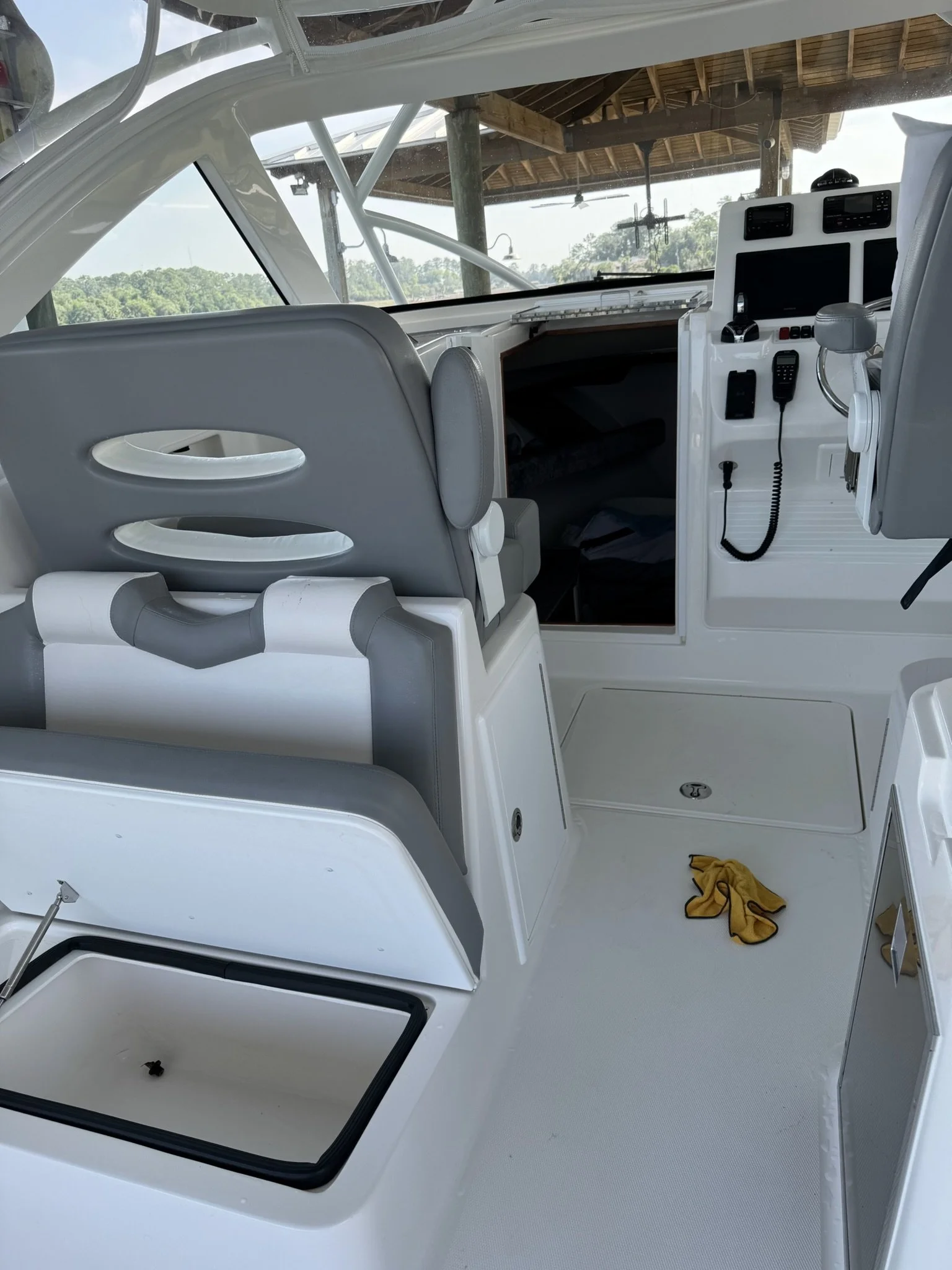 Interior of a boat cockpit with gray and white seats, a windshield, and various control panels. A yellow cloth is on the floor, with a wooden ceiling and trees visible through the windshield.