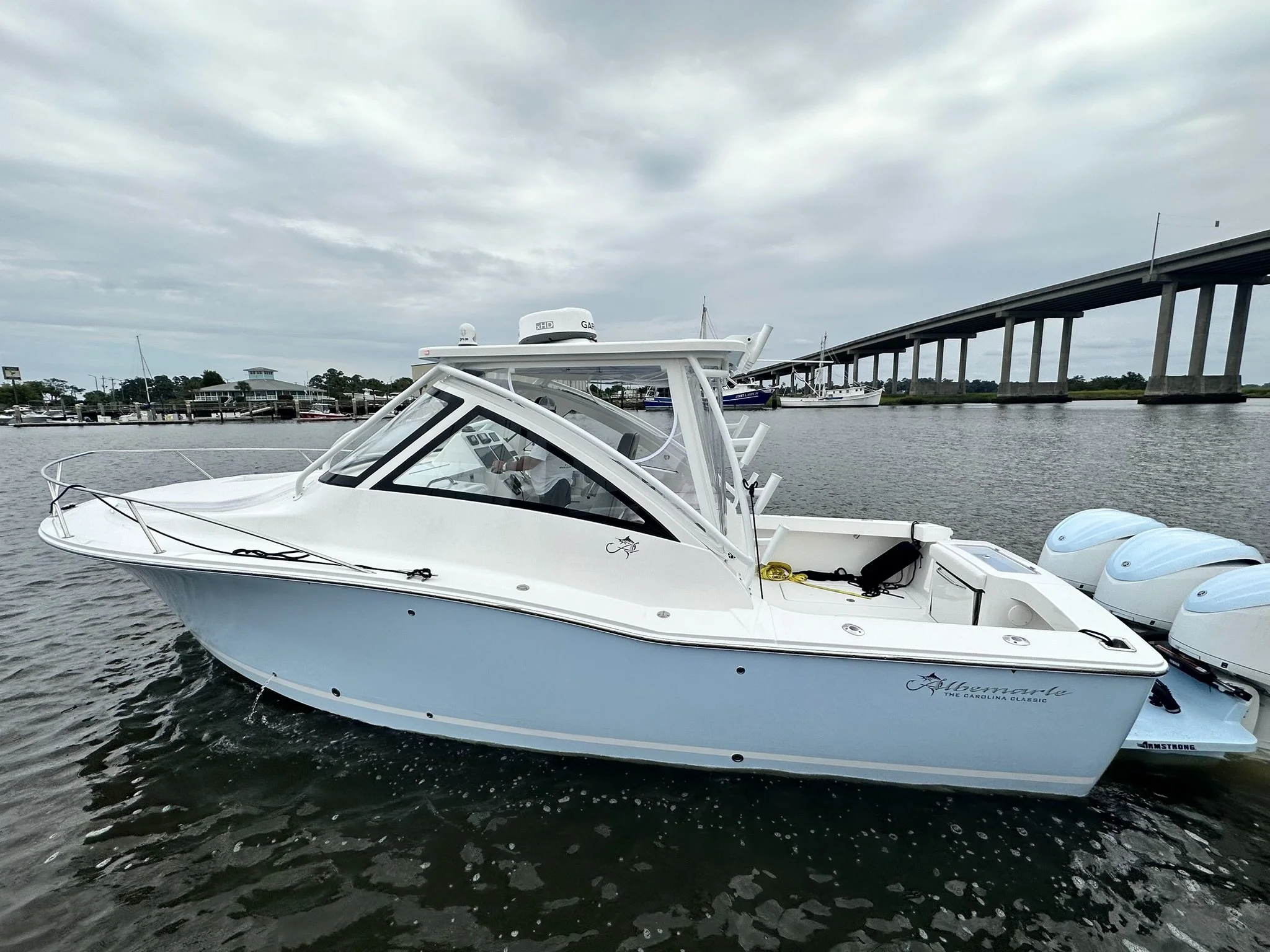 A white motorboat docked at a marina with other boats in the background under a cloudy sky, next to a large bridge.