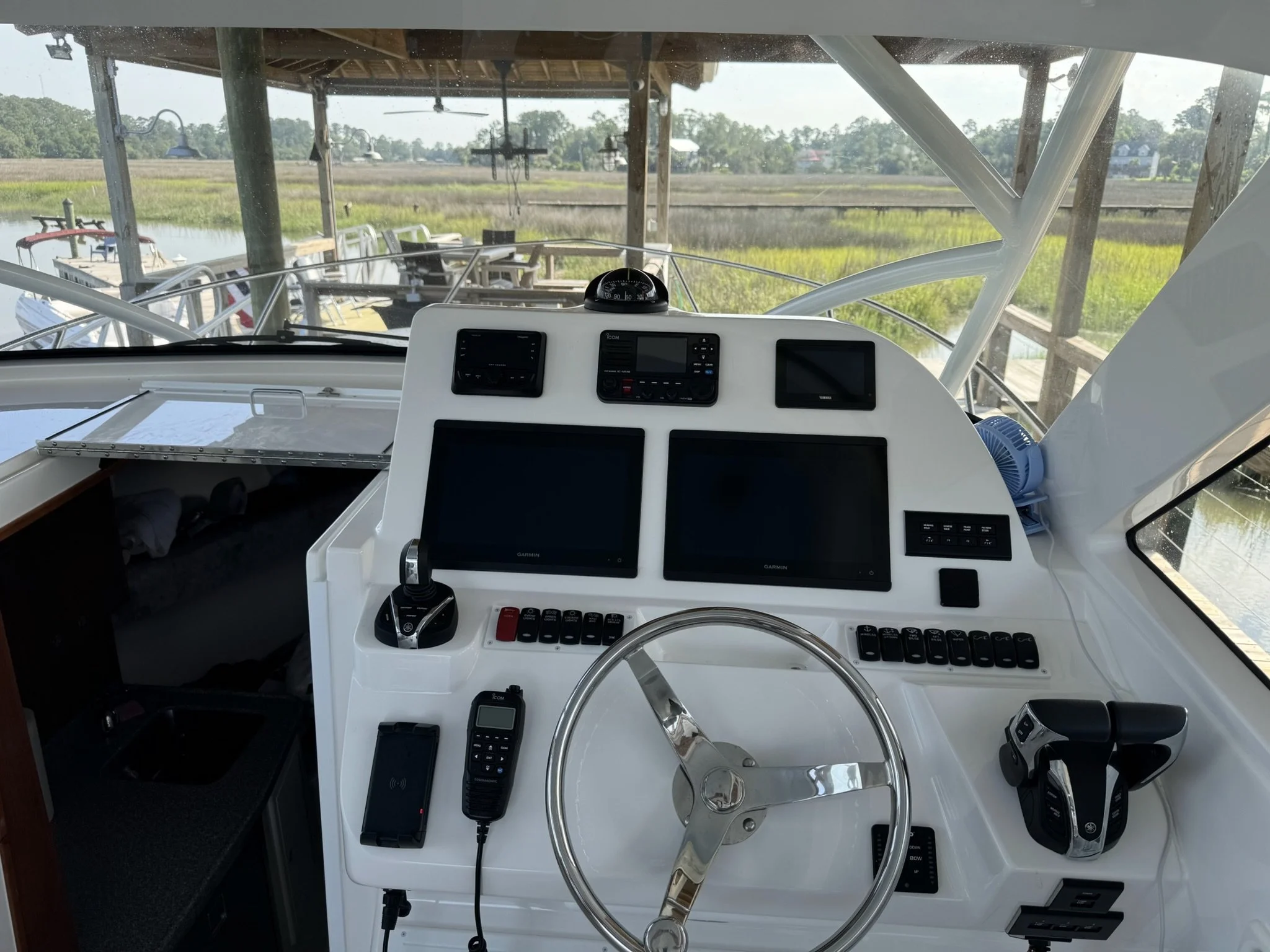 The image shows the interior of a boat's helm station with various navigation and communication equipment, including screens, radios, and control switches. The view from the helm looks out onto a dock with boats, a marshy waterway, and green landscap