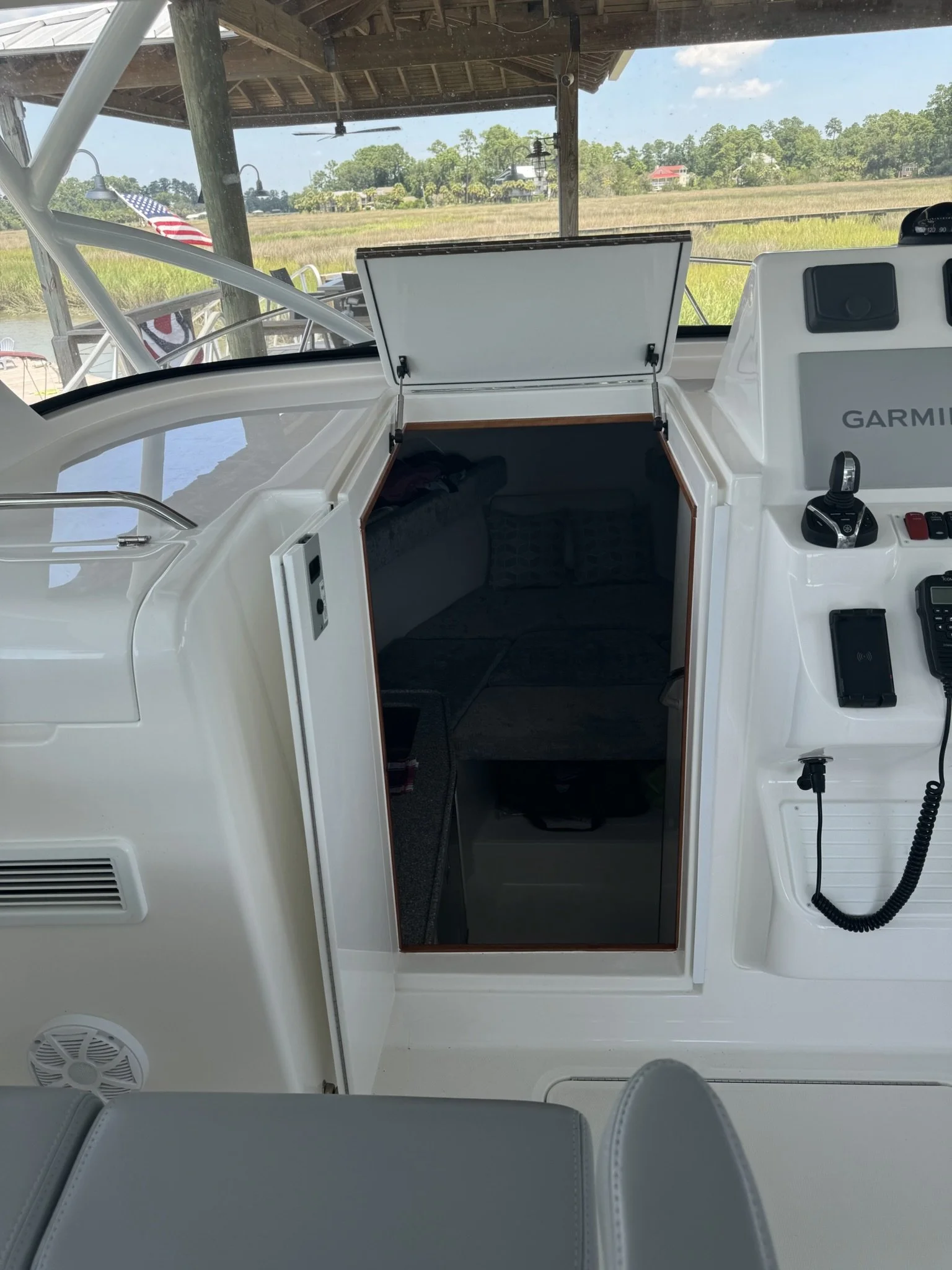 View from a boat showing an outdoor storage area with open hatch, part of the boat's interior, a cockpit with control devices, and a scenic landscape in the background with grass, trees, houses, and a blue sky.