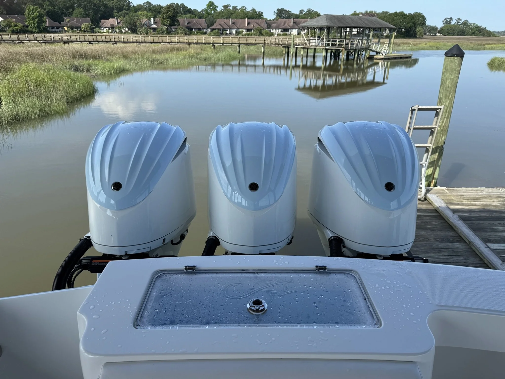 Four white outboard motors on the back of a boat, docked at a wooden pier on a calm waterway, with a covered boat lift and houses with brown roofs in the background.