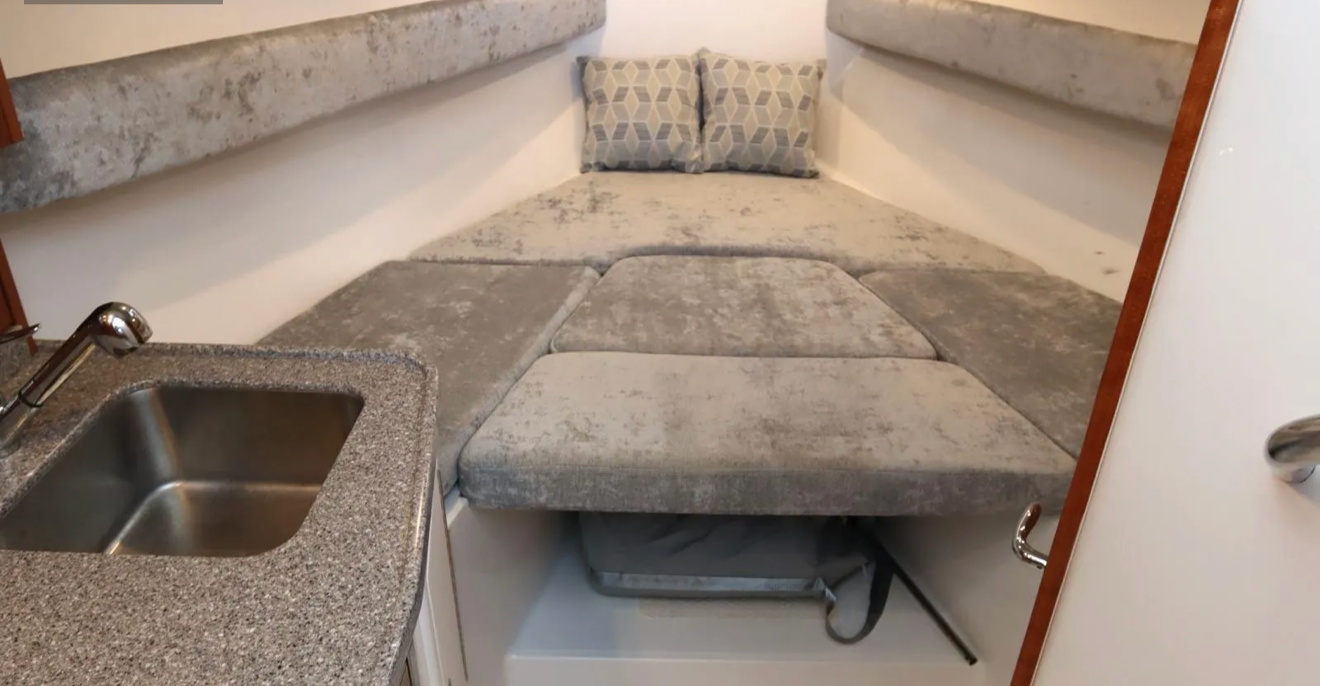 Small indoor space with built-in gray cushioned bench seating and pillows, a small sink with a speckled gray counter, and a white wall with cabinetry.
