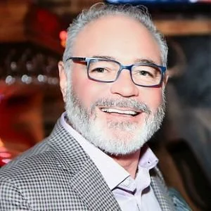 A smiling middle-aged man with gray hair and beard, wearing glasses and a gray checkered blazer, in an indoor setting with warm lighting.