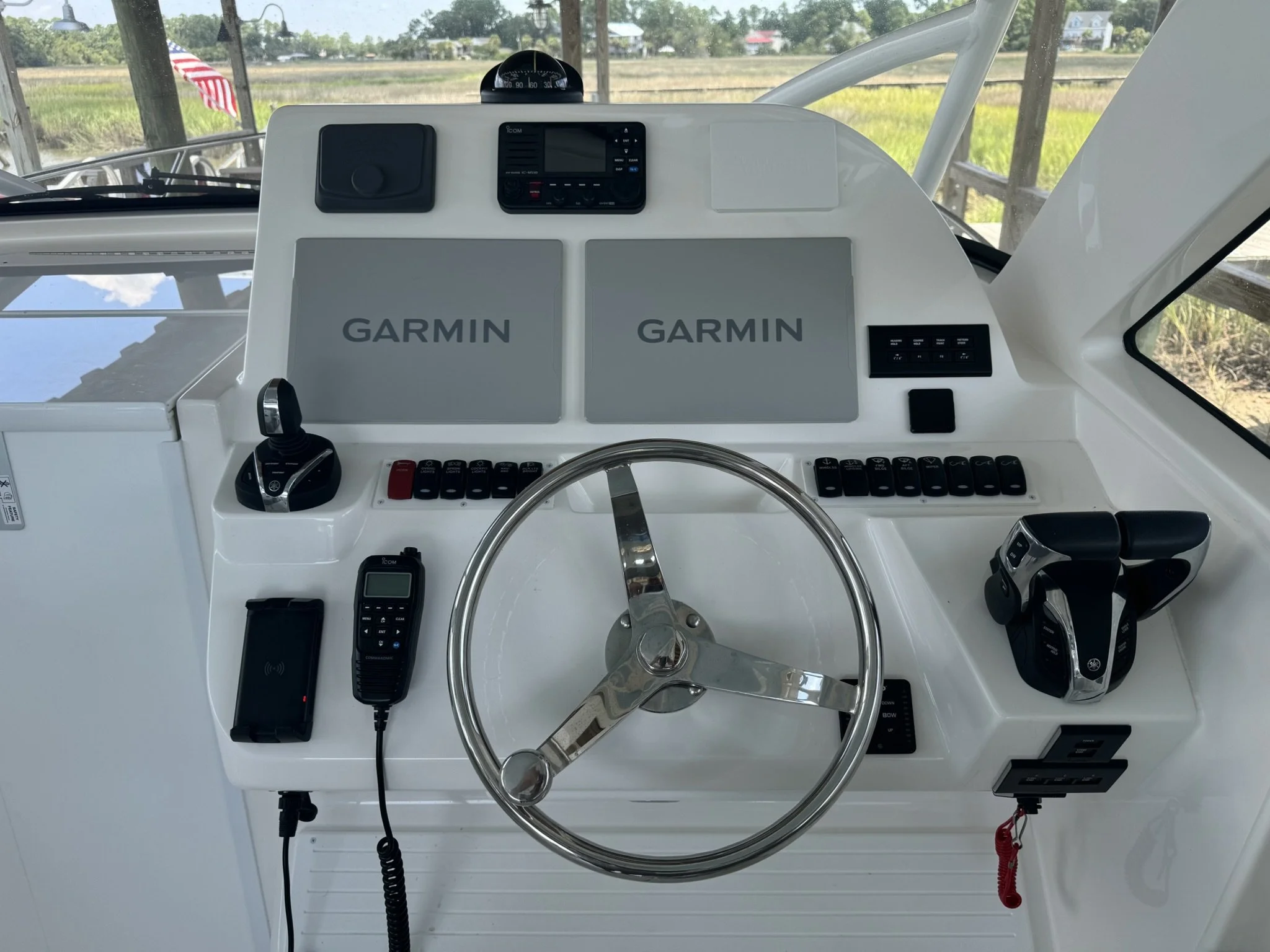 Boat helm station with navigation equipment, Garmin branded screens, a steering wheel, and various controls, with a view of the river and grassy fields outside.