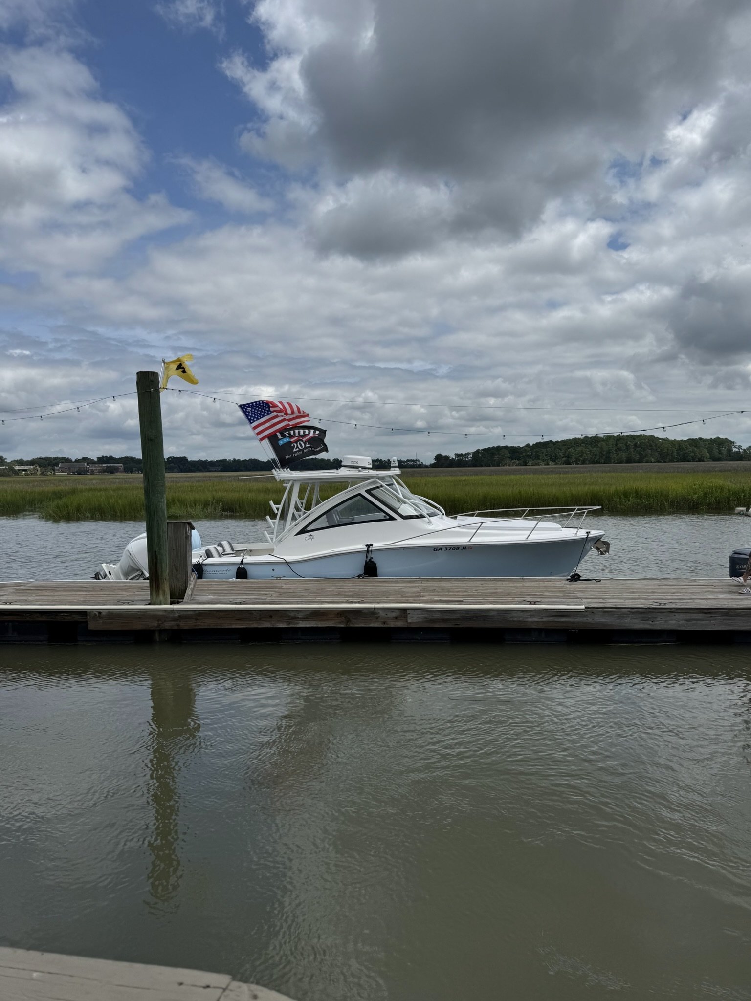 A white boat docked by a wooden pier on a calm body of water, with flags including an American flag and a Trump 2024 flag, under a cloudy sky with some blue patches, and greenery in the background.