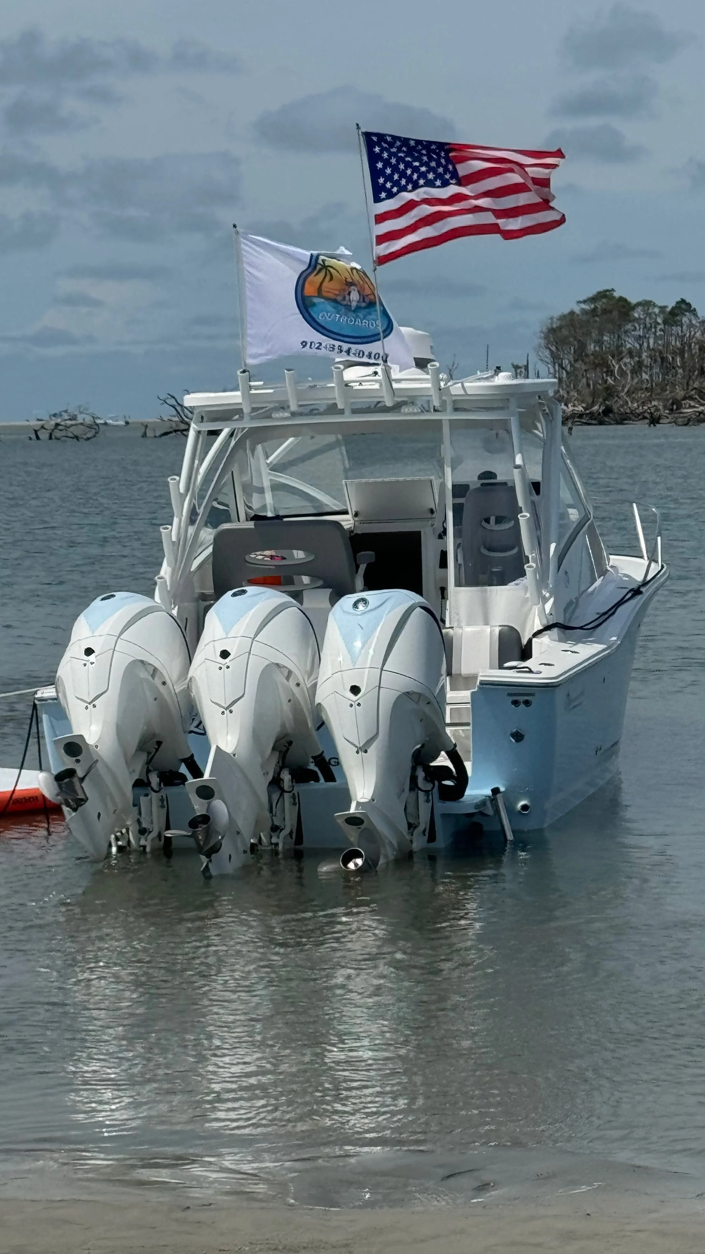 A boat with four outboard motors, American flag, and California state flag, anchored in water near a shoreline with trees and driftwood.