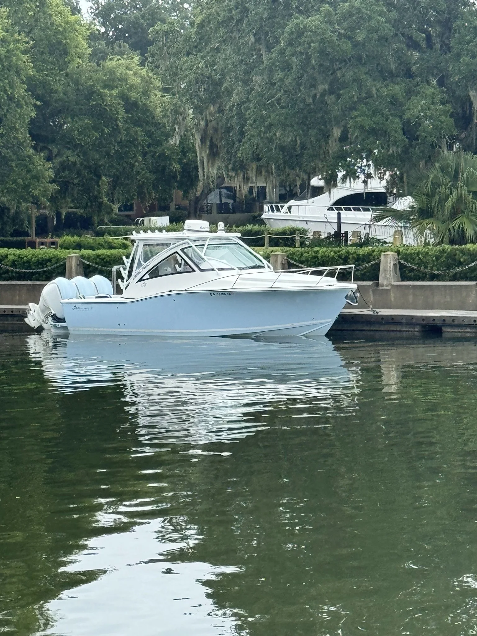 A white motorboat docked along a canal, with lush green trees and another boat in the background.